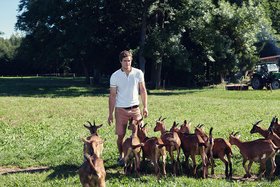 Ein junger Mann steht auf einer Wiese und wird von einer Gruppe brauner Ziegen umgeben.
