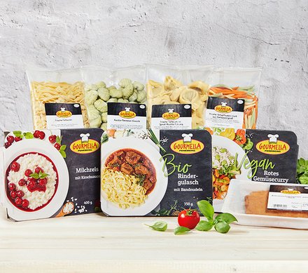 GOURMELLA-Produkte Eine Auswahl von GOURMELLA-Produkten, darunter verschiedene Teigwaren wie Tortellini, Gnocchi und Bandnudeln, sowie Fertiggerichte wie Milchreis mit Kirschsauce, Bio-Rindergulasch und veganes rotes Gemüsecurry, auf einem hellen Holztisch vor einer grauen Wand.