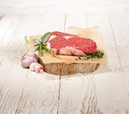 Frisches Steak auf Holzbrett Ein frisches Stück rohes Steak liegt auf Pergamentpapier, das auf einem rustikalen Holzstück präsentiert wird. Daneben liegen frischer Knoblauch, Rosmarin, Salbei und Oregano.